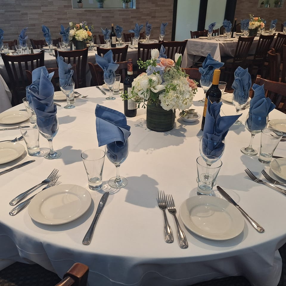 Elegantly set restaurant dining room with white tablecloths, blue napkins, floral centerpieces, wine bottles, and place settings.