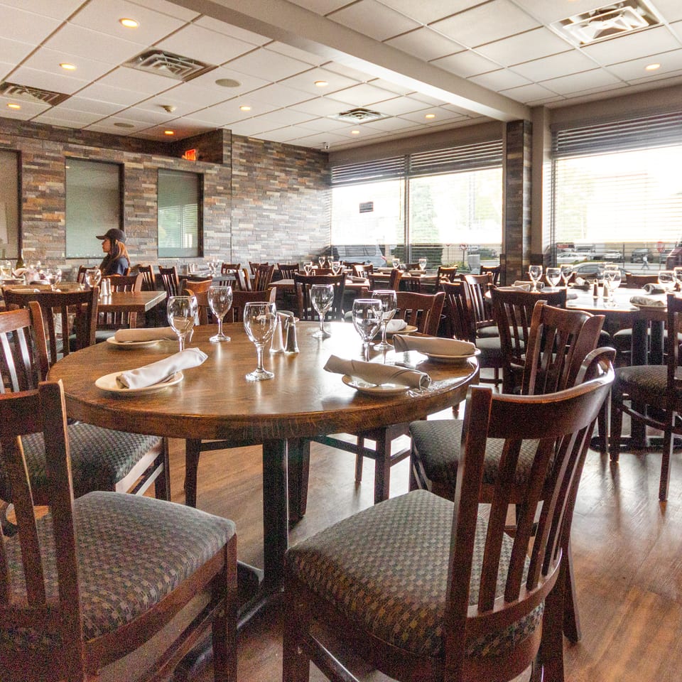 Bright restaurant dining room with wood tables, upholstered chairs, wine glasses, folded napkins, and large windows.