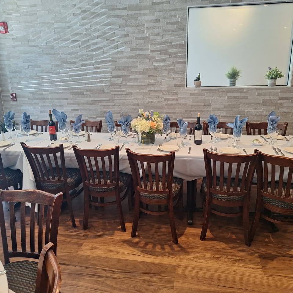 Elegant restaurant dining room with a long white-clothed table, blue napkins, wine bottles, floral centerpiece, and wooden chairs.