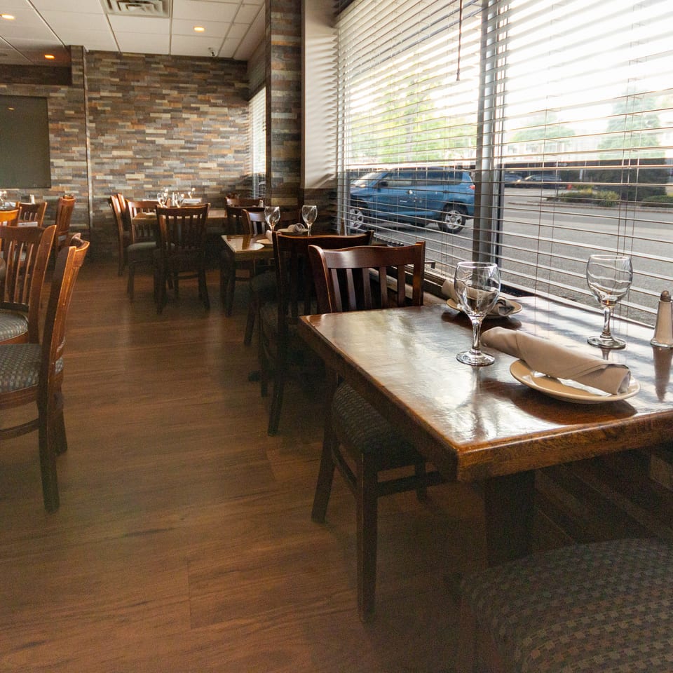 Warmly lit restaurant dining room with wood tables, set place settings, stone accent walls, and large windows overlooking the street.