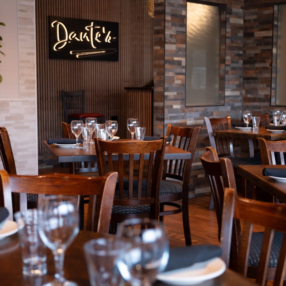 Warmly lit restaurant dining room with wooden tables, upholstered chairs, place settings, wine glasses, and a glowing wall sign.