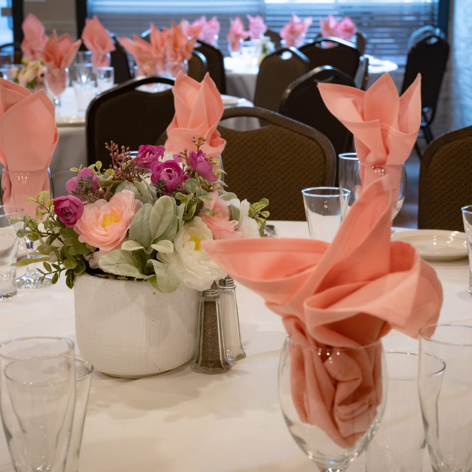 Restaurant dining room tables decorated with pink folded napkins, glassware, white linens, and colorful floral centerpieces.