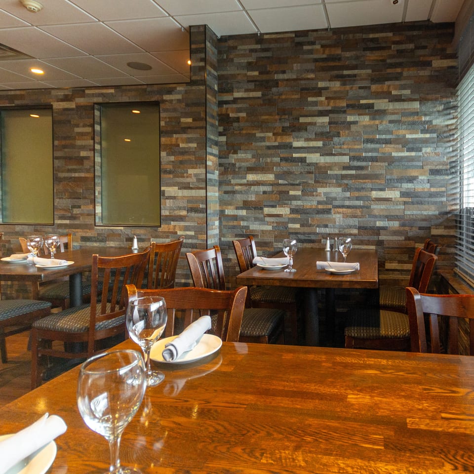 Warmly lit restaurant dining room with wooden tables, set plates, wine glasses, upholstered chairs, and a stacked-stone accent wall.