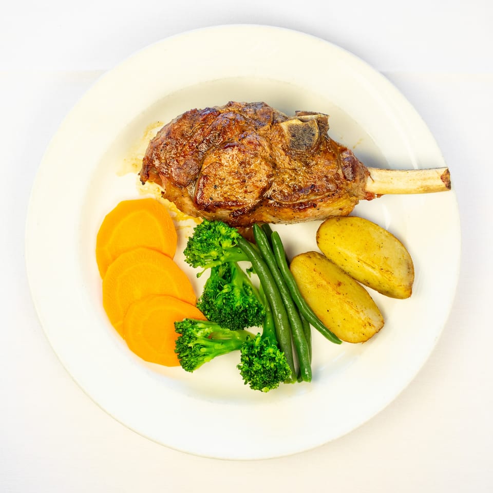 Grilled bone-in steak served with roasted potatoes, sliced carrots, green beans, and broccoli on a white plate.