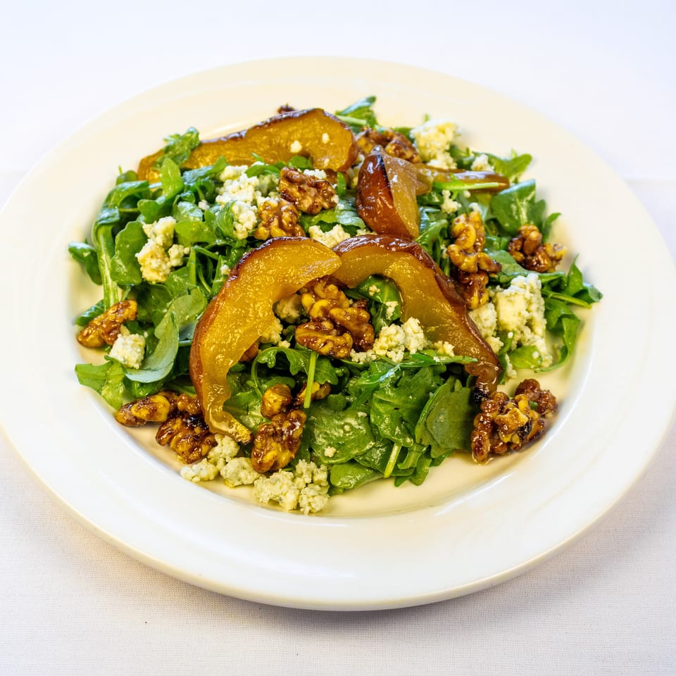 Green salad topped with roasted pear slices, candied walnuts, and crumbled blue cheese on a white plate.